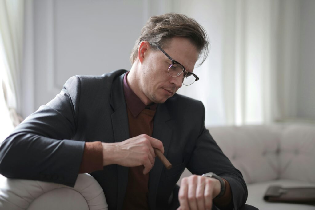 Man in a suit, checking time on wristwatch while holding a cigar indoors.
